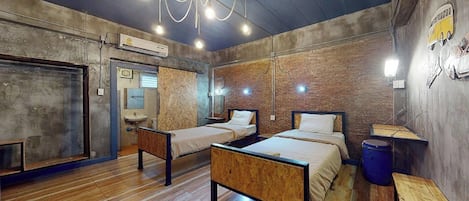 Una habitación con dos camas, piso de madera, pared de ladrillo y techo con tuberías a la vista.