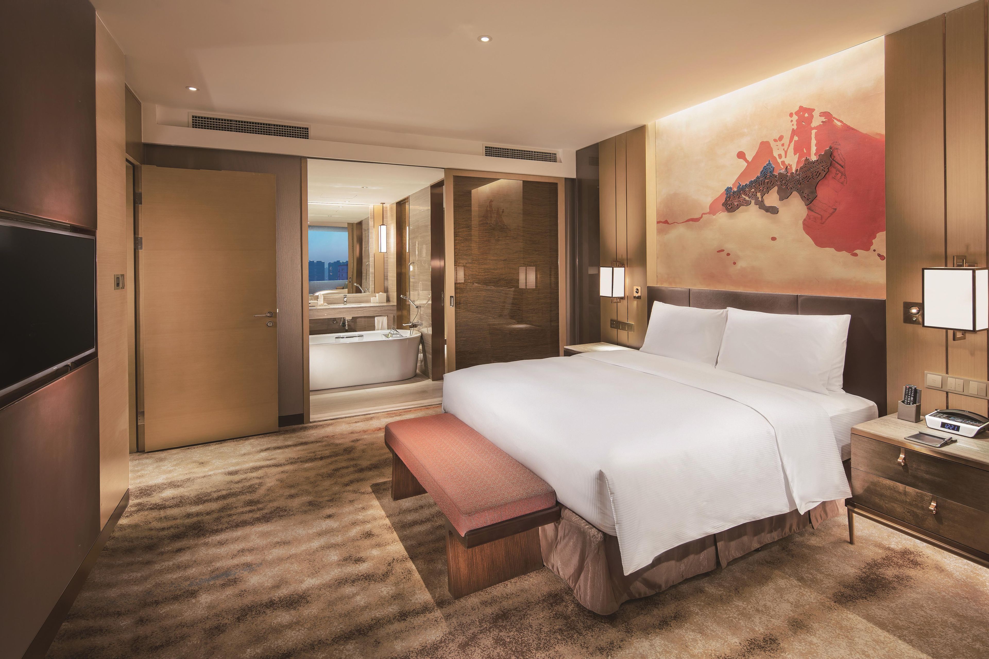 Habitación de hotel con una cama grande, un banco, un baño con bañera y un televisor de pared.
