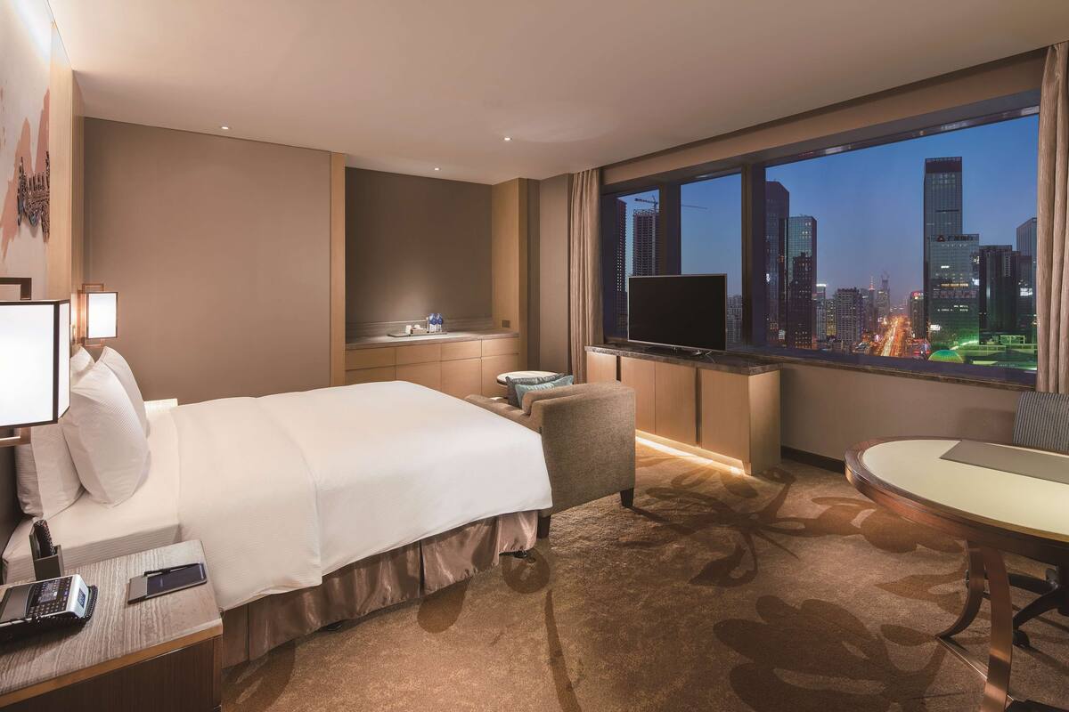 A hotel room with a bed, a desk, a chair, a TV, and a view of a cityscape at dusk.