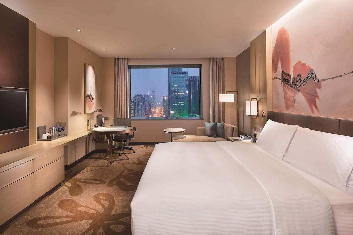 A hotel room with a large bed, a desk, a chair, a small table, a TV, and a window with a city view.