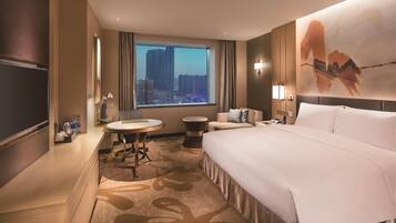 A hotel room with a large bed, a desk, a chair, a round table, a sofa, a TV, and a window with a city view.