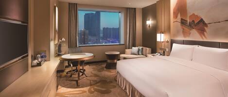 A hotel room with a large bed, a desk, a chair, a round table, a sofa, a TV, and a window with a city view.
