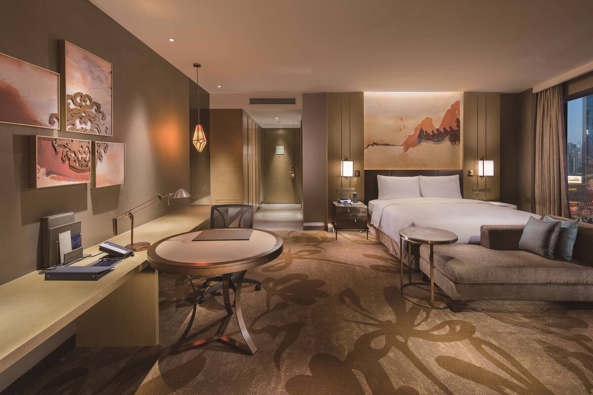 A hotel room with a bed, a sofa, a desk, and a chair. There is a painting on the wall and a lamp on the nightstand.