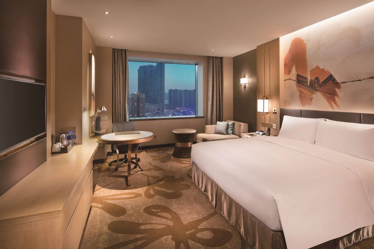 A modern hotel room with a large bed, a desk, a round table, and a view of a cityscape through a large window.