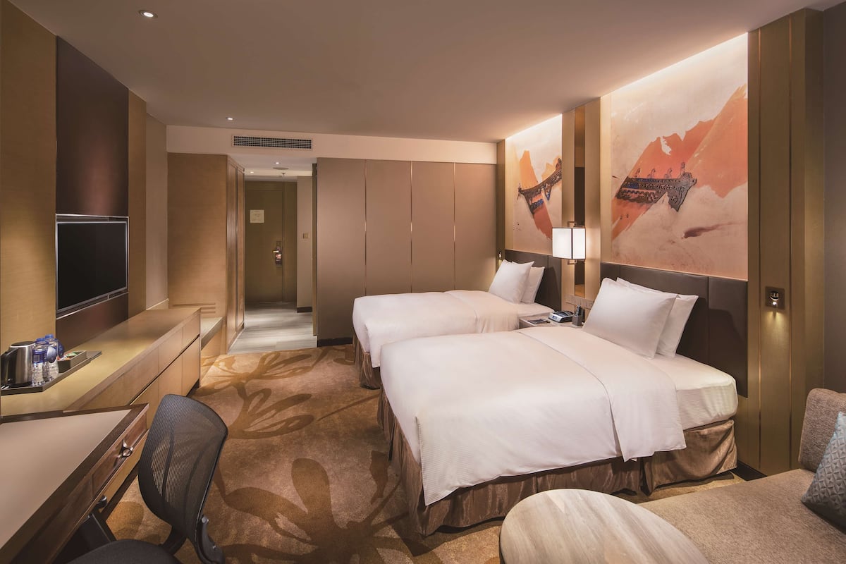 A hotel room with two beds, a desk with a chair, a TV, and a large wall mural.