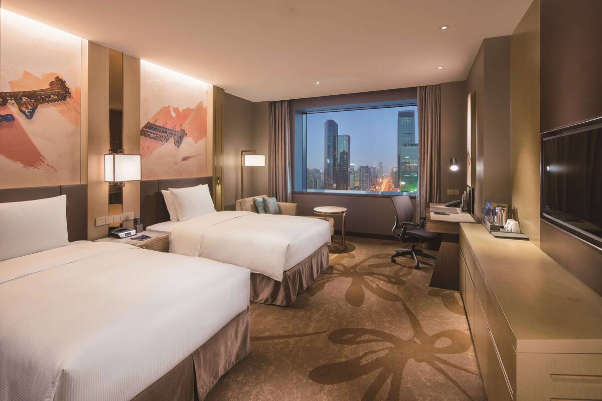 A hotel room with two beds, a desk, a chair, a TV, and a large window with a city view.