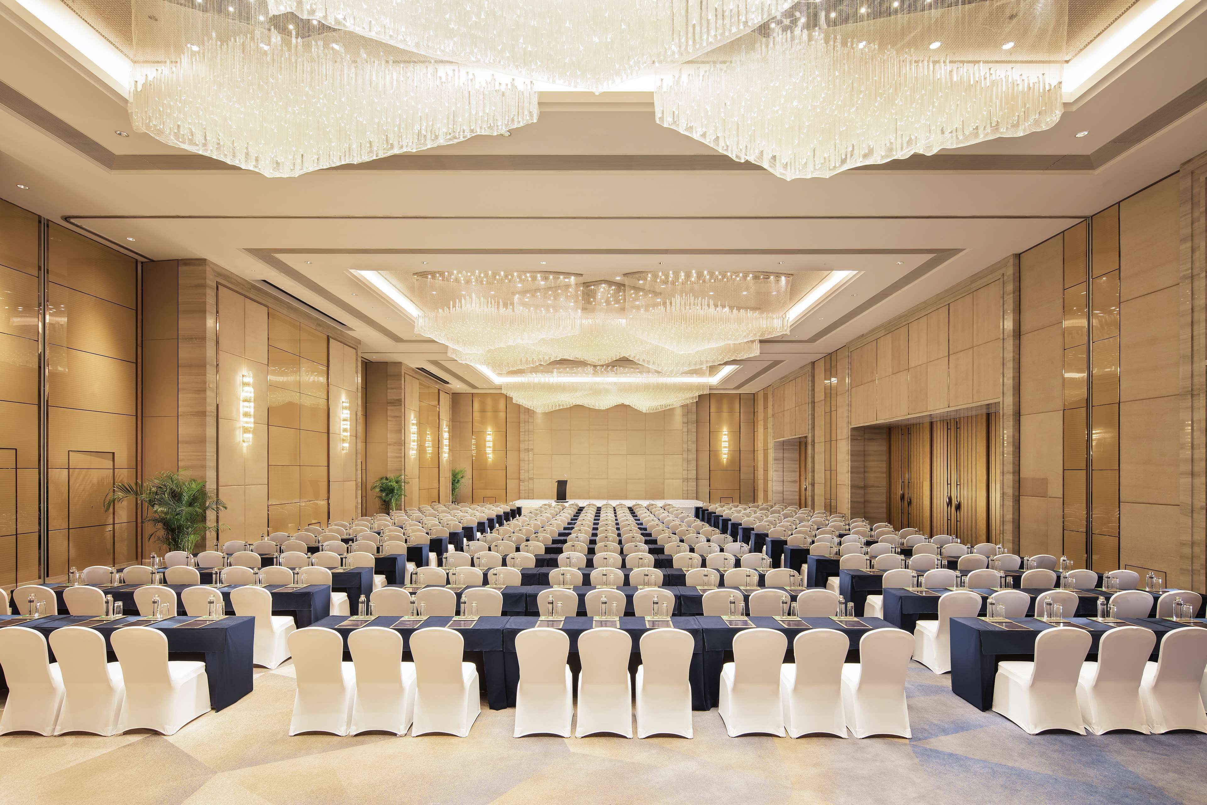 A large conference room with rows of tables and chairs, a chandelier, and a podium at the front.