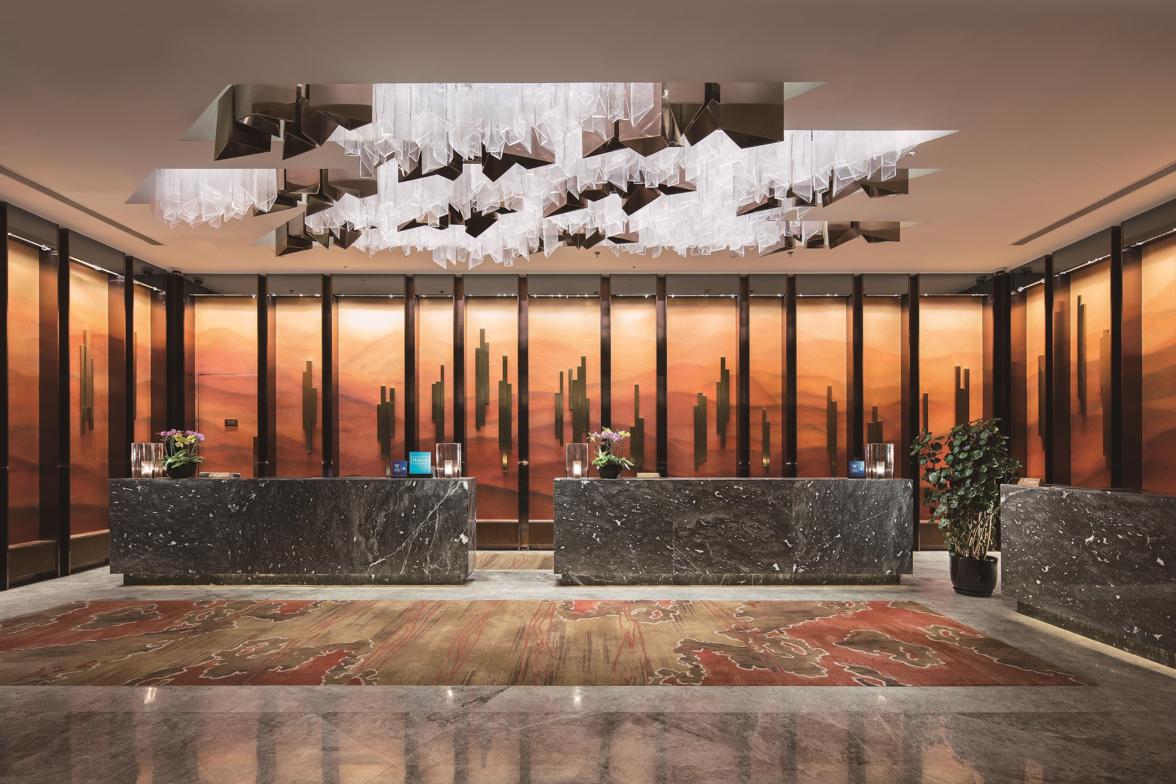 Zona de recepción de hotel con diseño moderno, destacando un mostrador de mármol, una gran instalación de luces abstractas y un mural con vistas a la ciudad.