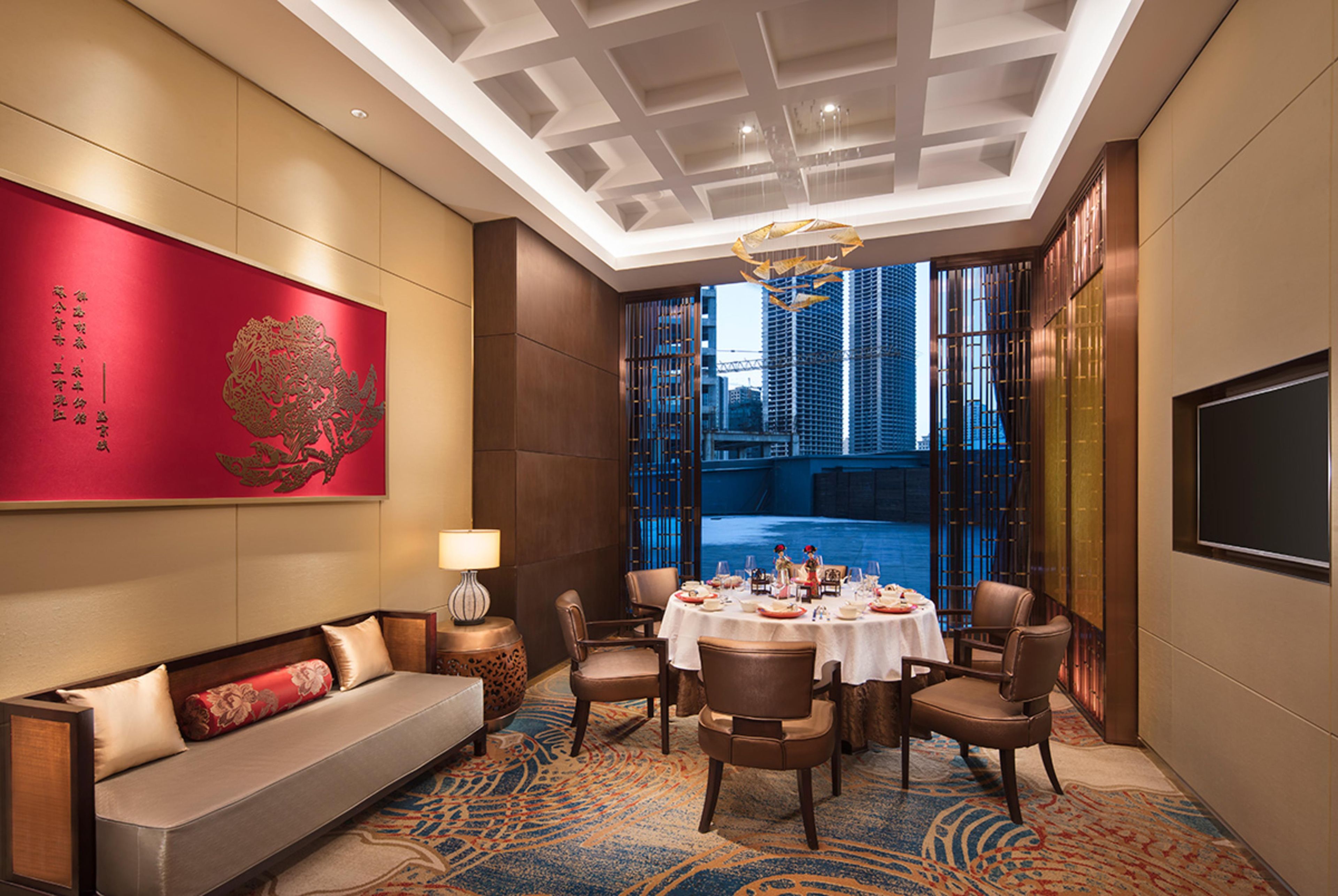A hotel room with a sofa, dining table, chairs, a TV, a large red artwork, and a view of a cityscape.