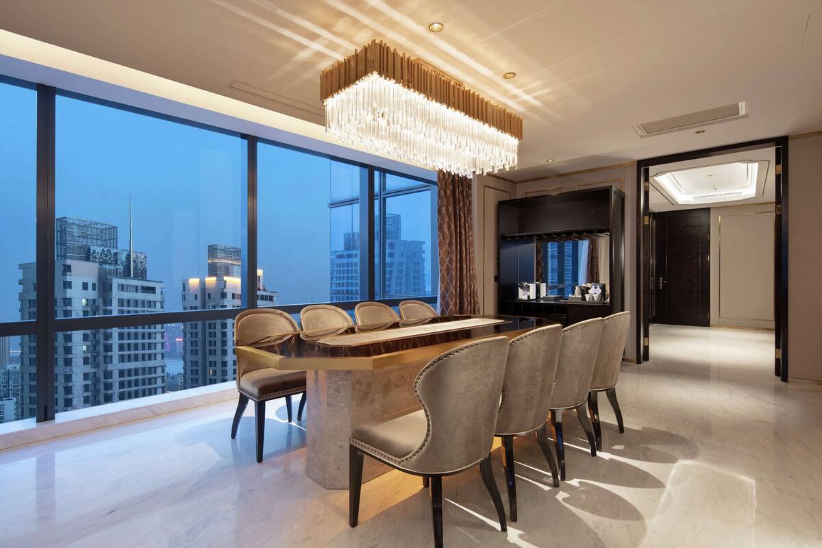 A modern dining area with a large table and chairs, a chandelier, and a city view through large windows.