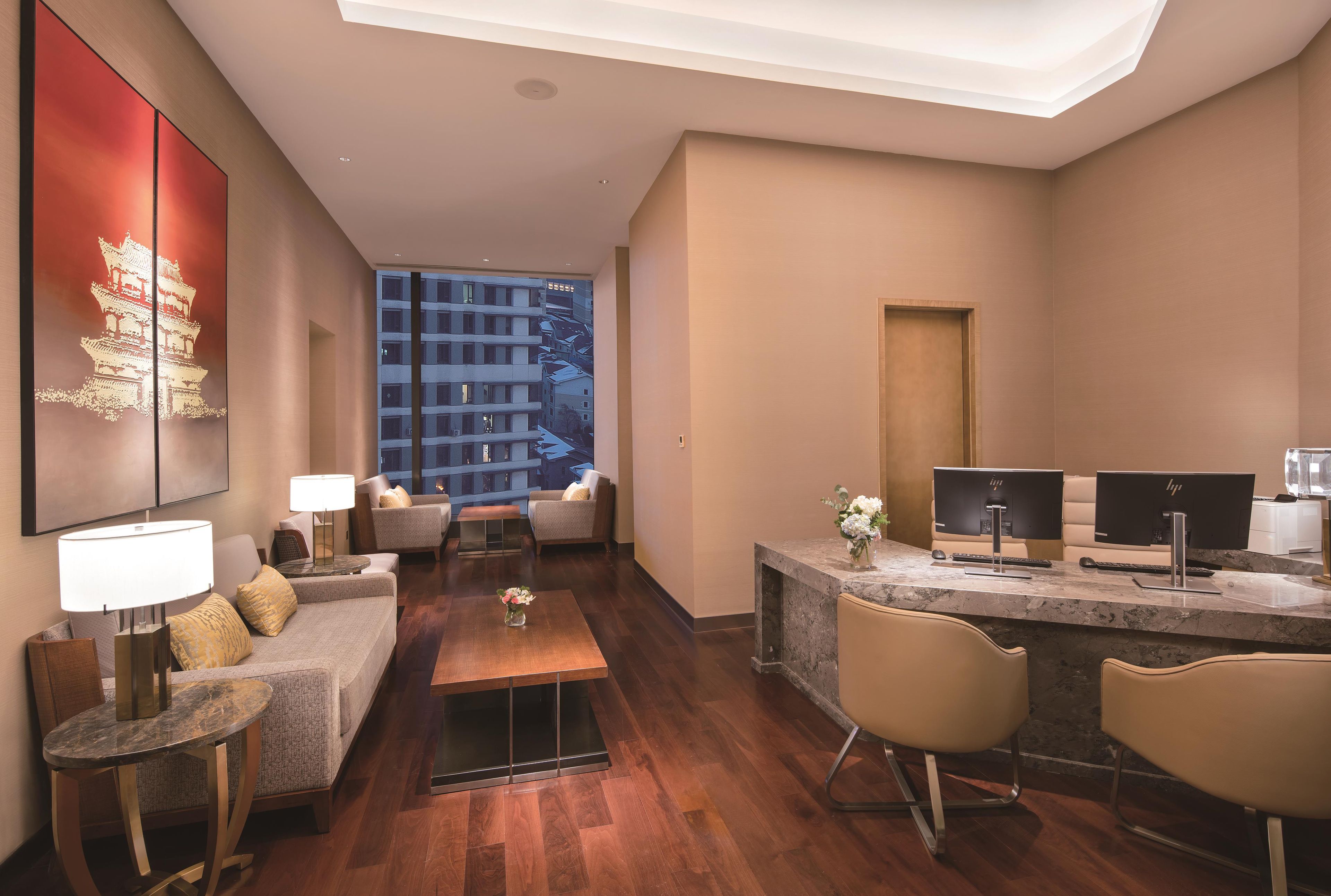 A modern hotel room with a living area, a desk with computers, and a large window overlooking a cityscape.