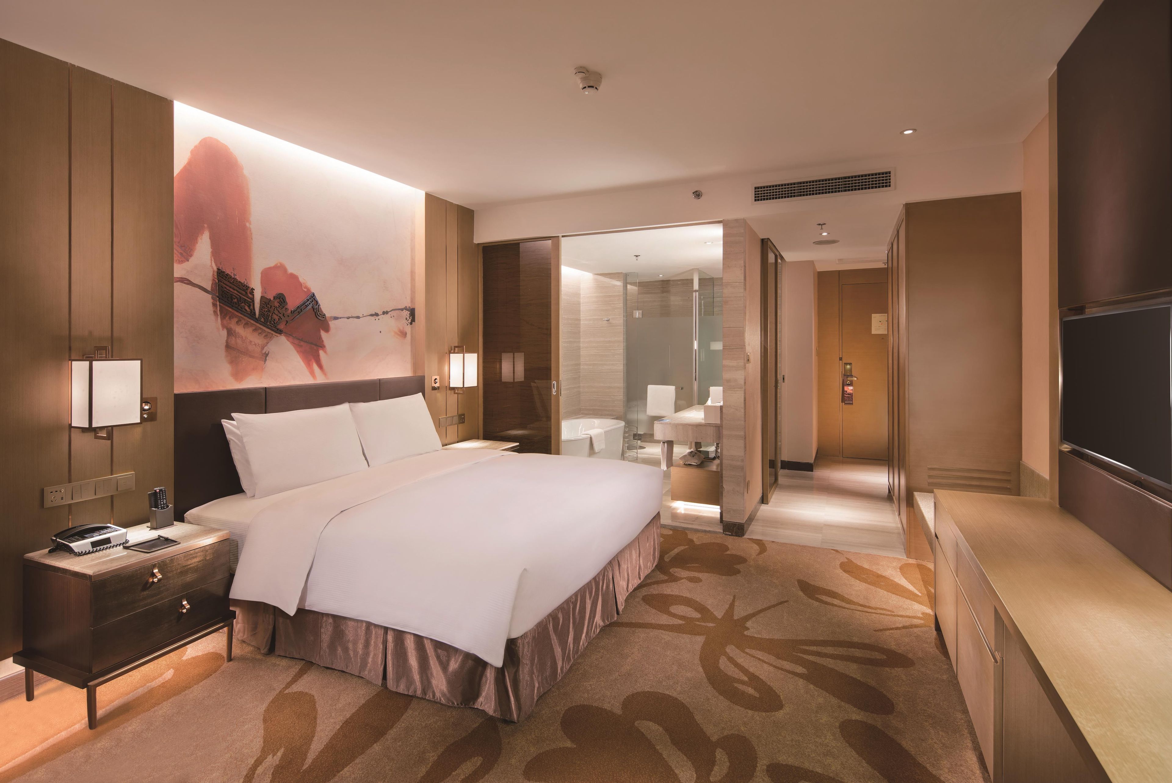 Ein Hotelzimmer mit einem großen Bett, Nachttischen, einem Schreibtisch, einem Sessel und einem Fernseher. Ein Badezimmer ist durch eine offene Tür sichtbar.