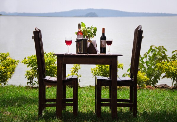 Mesa de madera con dos sillas y una botella de vino, sobre un césped con un lago y montañas al fondo.