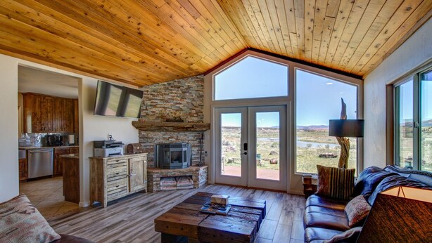 A spacious living area with a stone fireplace, wooden ceiling, and large windows offering a view of the outdoors. - Sevier River Retreat (Hatch)
