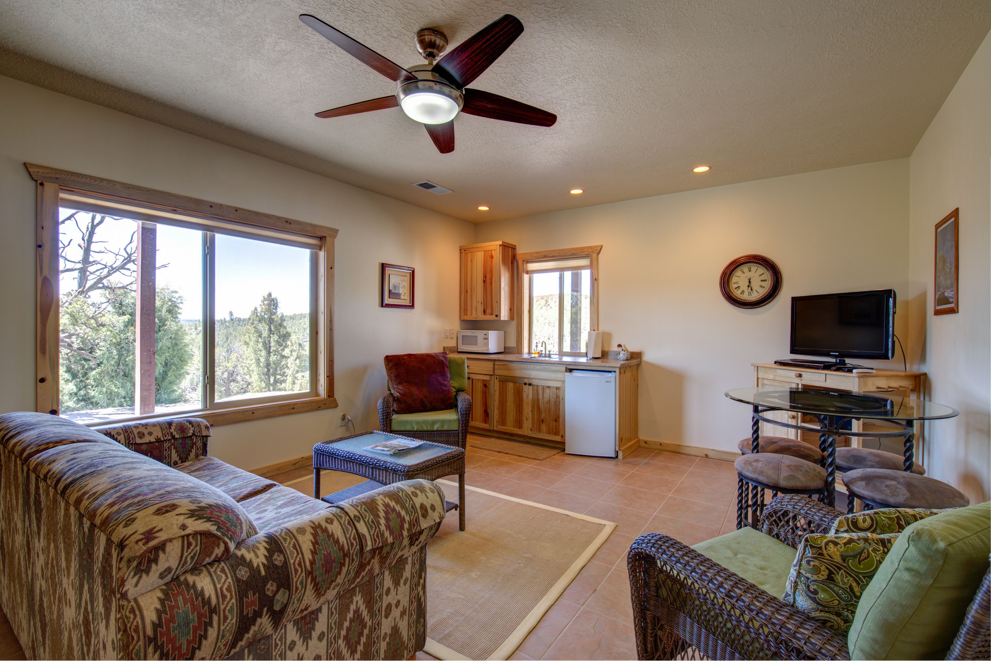 A spacious living room with a large window, a ceiling fan, a kitchen area, and a television.