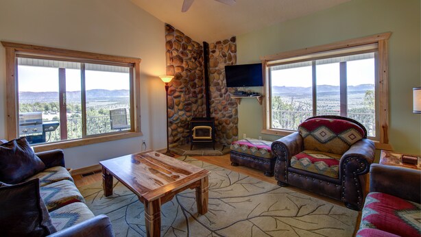 A cozy living room with a stone fireplace, large windows offering a view of the mountains, a ceiling fan, and a television mounted on the wall. - Ridgetop Hideaway (Hatch)