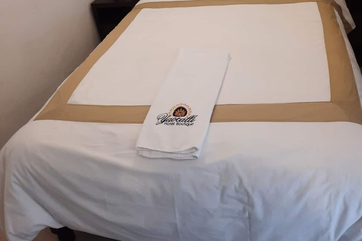 A neatly made bed with a white and beige bedspread, a folded towel with a logo, and a dark headboard.