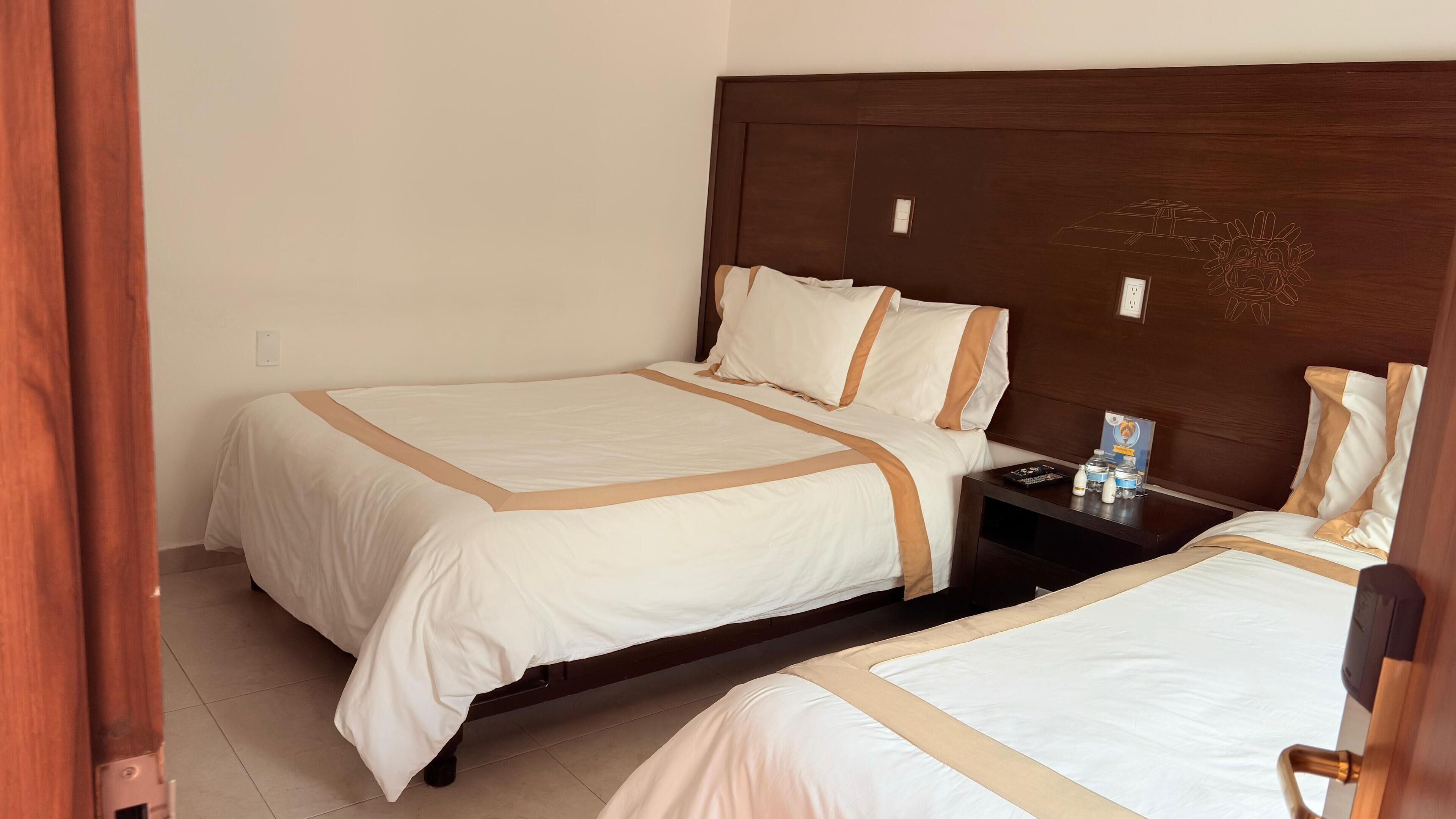 A hotel room with two beds, each with white bedding and brown trim. There is a wooden headboard, a nightstand with a lamp, and a small table with a water bottle and a book.