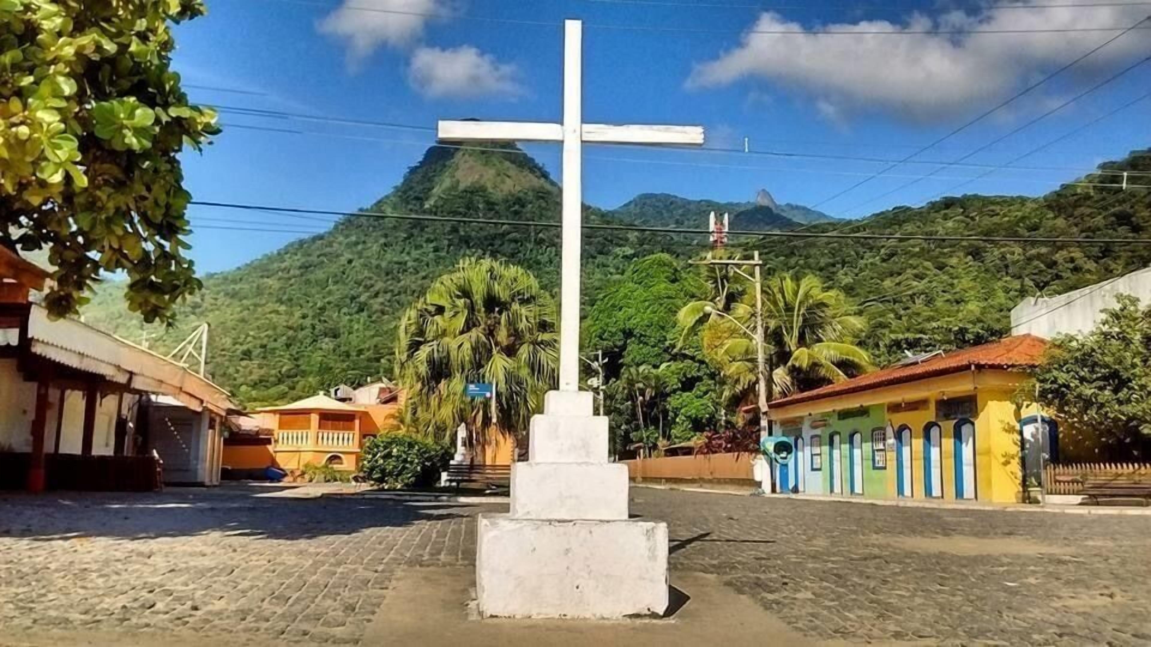 A cross monument in a town square with buildings and mountains in the background.