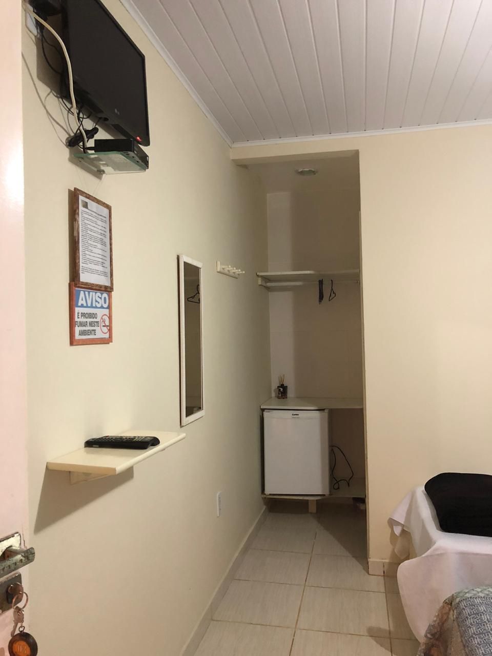 Una habitación pequeña y estrecha con techo blanco, televisor montado, refrigerador pequeño, espejo y un aviso en la pared.