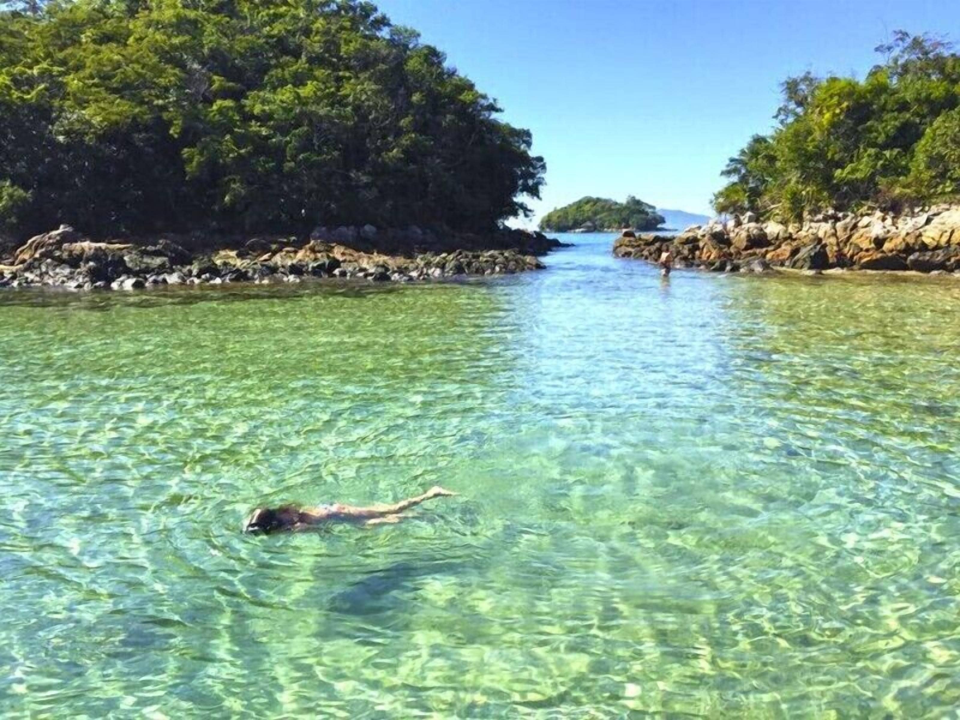 A swimmer in clear water with rocky shores and dense greenery.