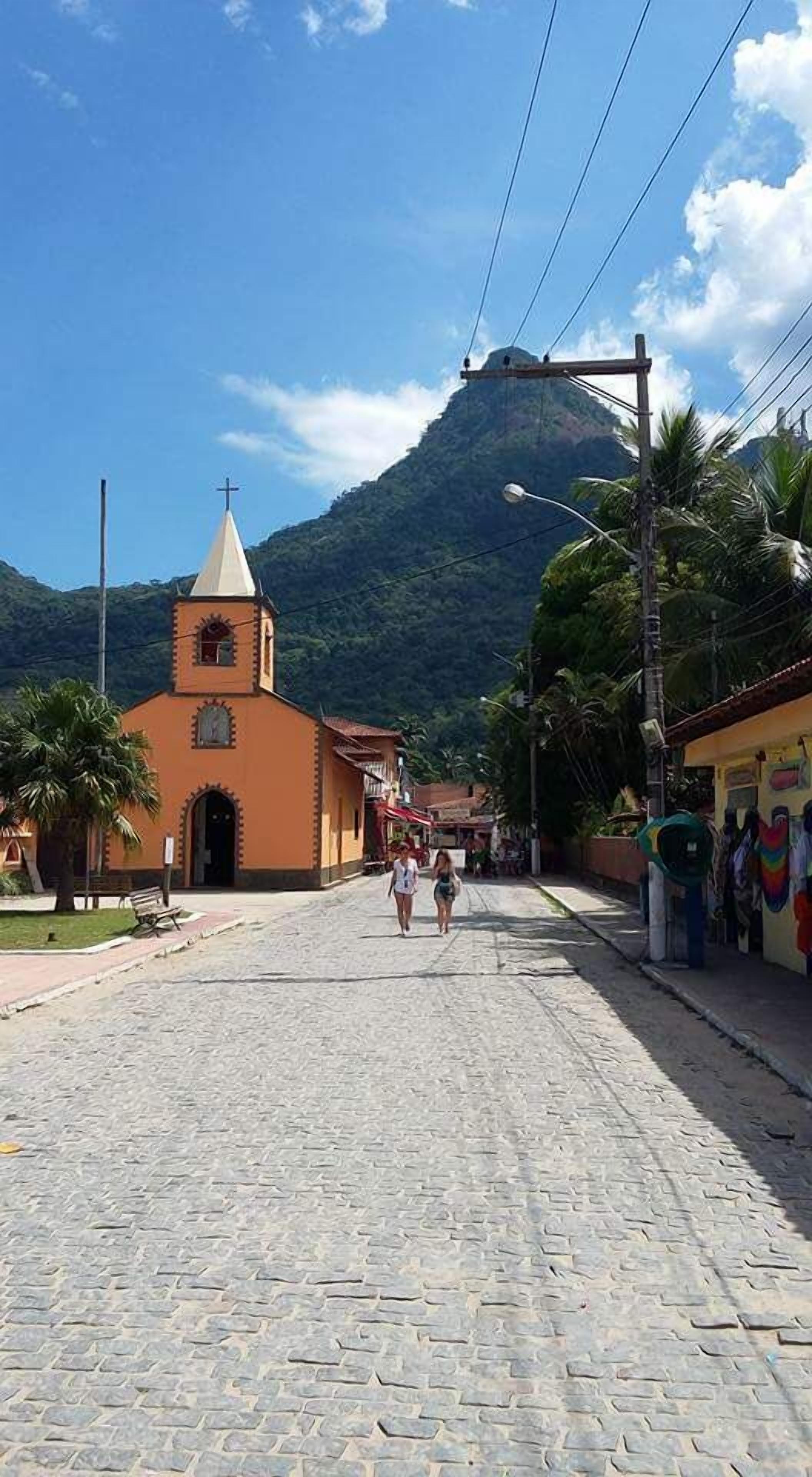 A cobblestone street leading to a church with a bell tower, surrounded by greenery and palm trees.
