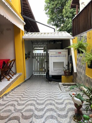 A narrow outdoor corridor with tiled flooring, a white gate, a white air conditioning unit, and a small table with chairs. - Recanto da Ana Alice (Angra dos Reis)