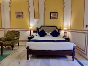 A bed with a wooden frame, two bedside tables with lamps, a floral armchair, and two framed pictures on the wall. - Narain Niwas Palace (Jaipur)