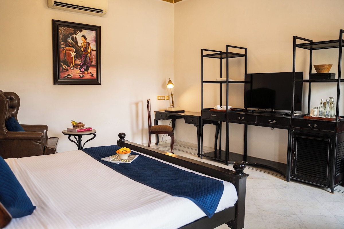 Habitación de hotel con cama, escritorio con televisor, silla, mesita con lámpara y un cuadro en la pared.