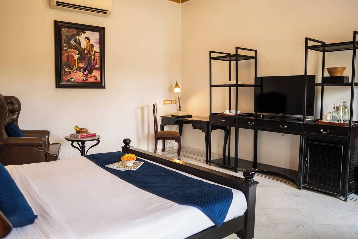 Une chambre d’hôtel avec un lit, un bureau avec une télévision, une chaise, une petite table avec une lampe et un tableau au mur.
