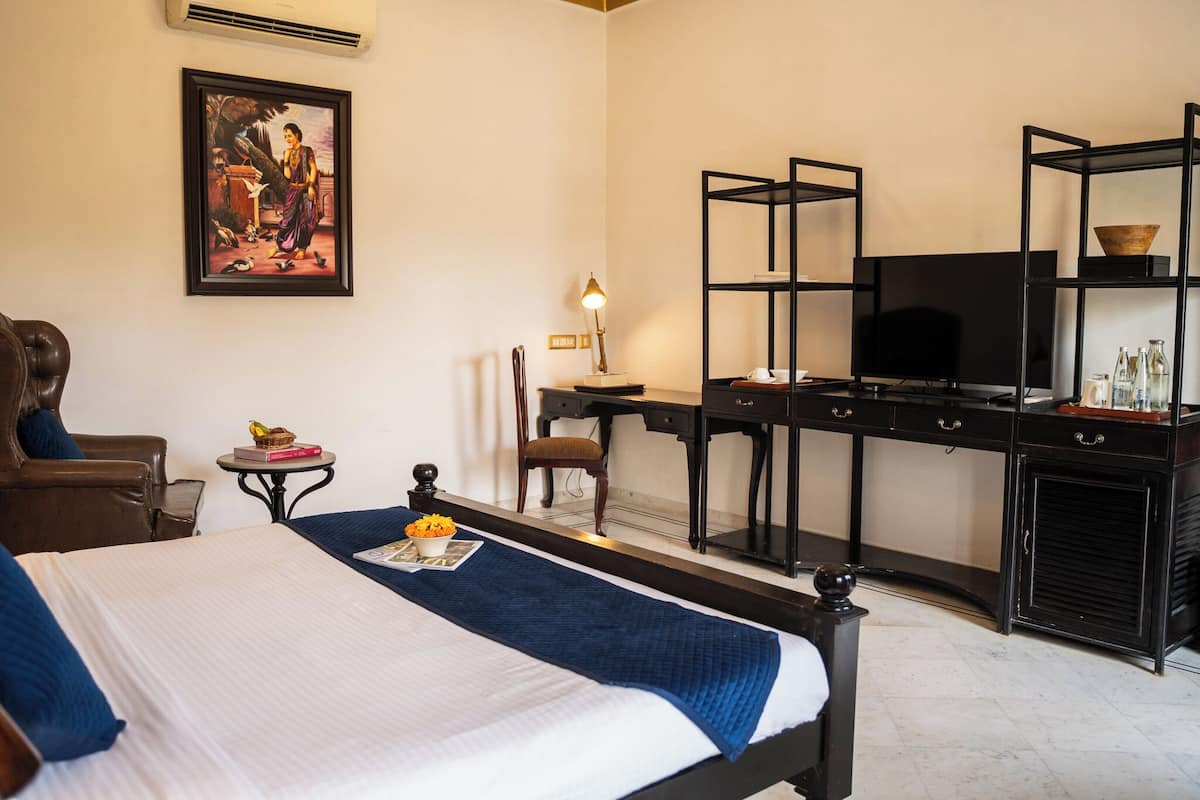 A hotel room with a bed, a desk with a TV, a chair, a small table with a lamp, and a painting on the wall.