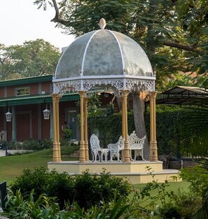 Ein Gazebo mit Kuppeldach und verzierten Geländern, umgeben von Grünpflanzen und einem Gebäude mit einem Gründach.