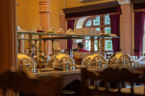 A breakfast buffet with glass display cases and golden domed covers, a person in the background, and a window with curtains.
