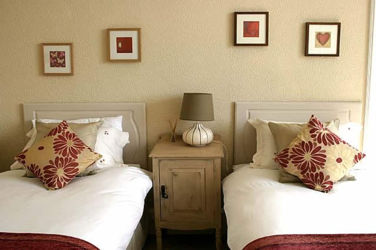 Ein Hotelzimmer mit zwei Betten, jeweils mit weißer Bettwäsche und roten Kissen mit Blumenmuster. Ein kleiner Nachttisch mit Lampe und dekorativer Vase ist vorhanden. Die Wände sind beige und mit gerahmten Bildern versehen.