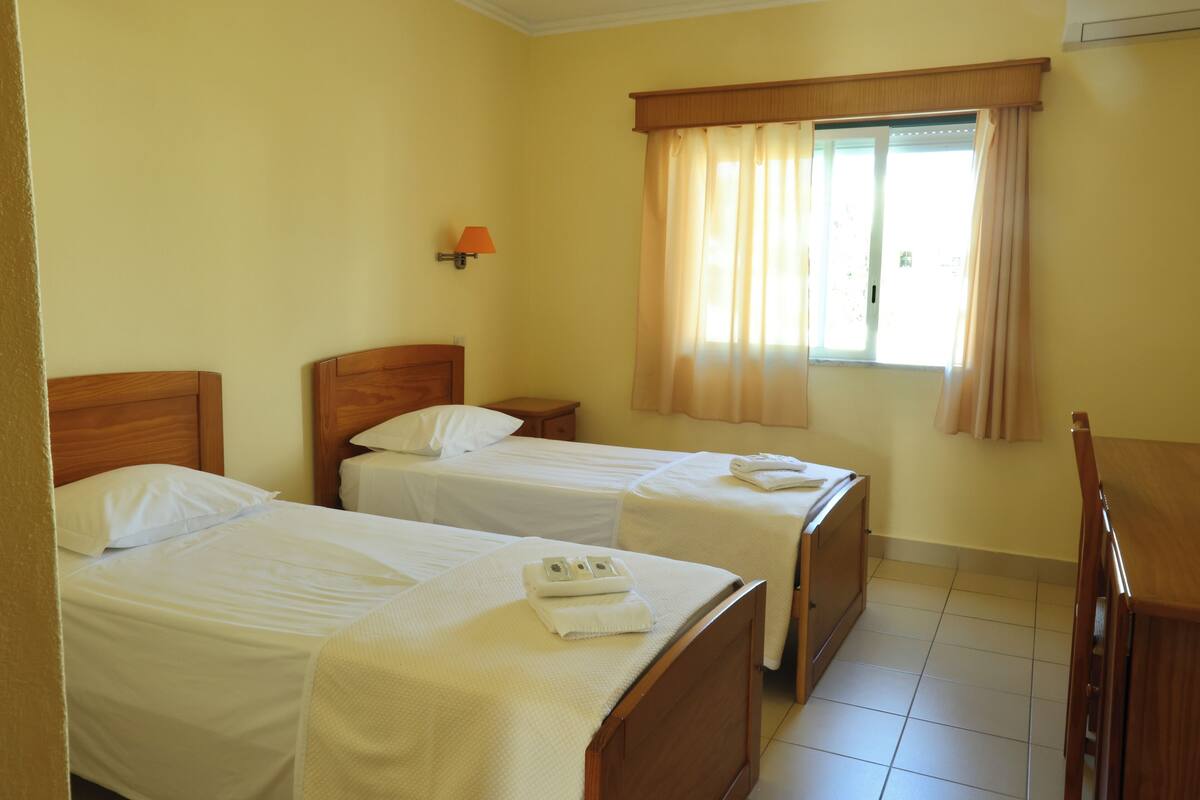 A hotel room with two single beds, a wooden headboard, a nightstand, a table, and a window with curtains.