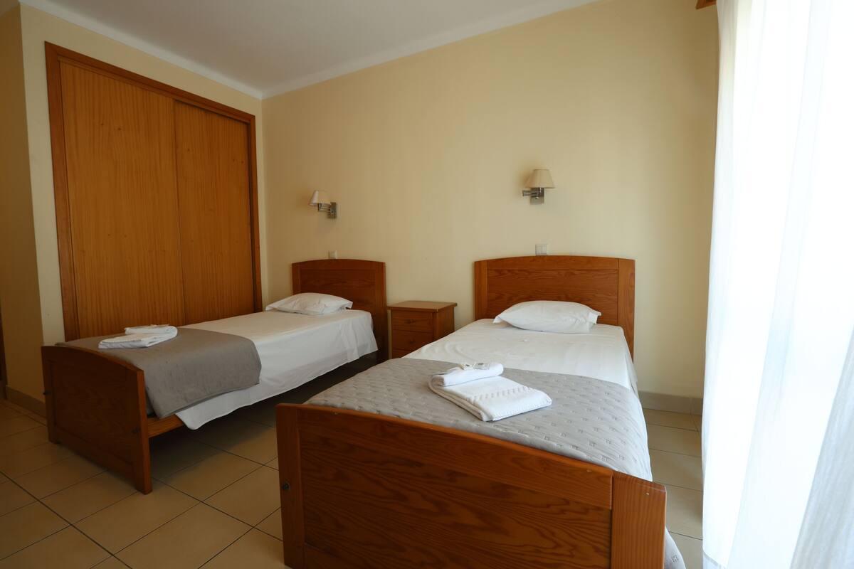 A hotel room with two wooden beds, a wardrobe, bedside tables, and wall-mounted lamps.