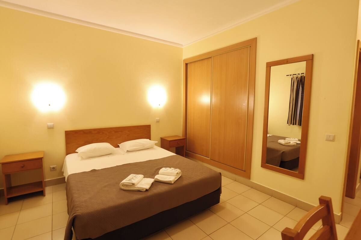 A hotel room with a bed, two bedside tables, a wardrobe, a mirror, and a chair.