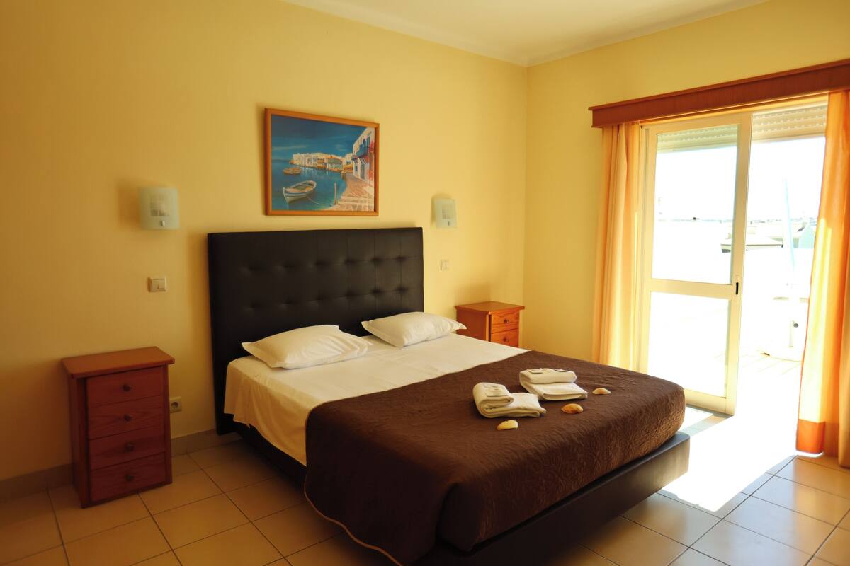 A hotel room with a bed, a nightstand, a dresser, a painting on the wall, and a window with curtains.