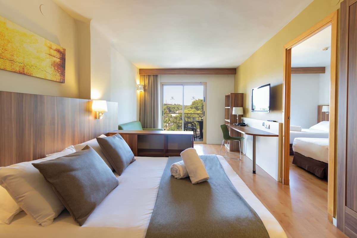 A hotel room with a bed, a desk, a TV, and a balcony with a view of trees.