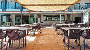Un patio exterior con varias mesas y sillas, una zona de estar cubierta y vista al interior del edificio.