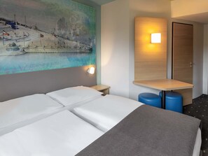 Habitación de hotel con cama, mesita de noche, lámpara de pared, espejo y un cuadro con una escena de playa.