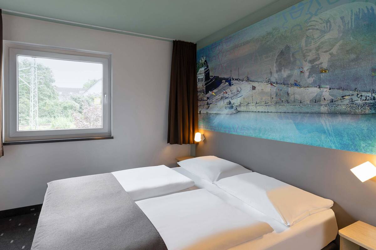A hotel room with a bed, a window, a painting, and a nightstand.
