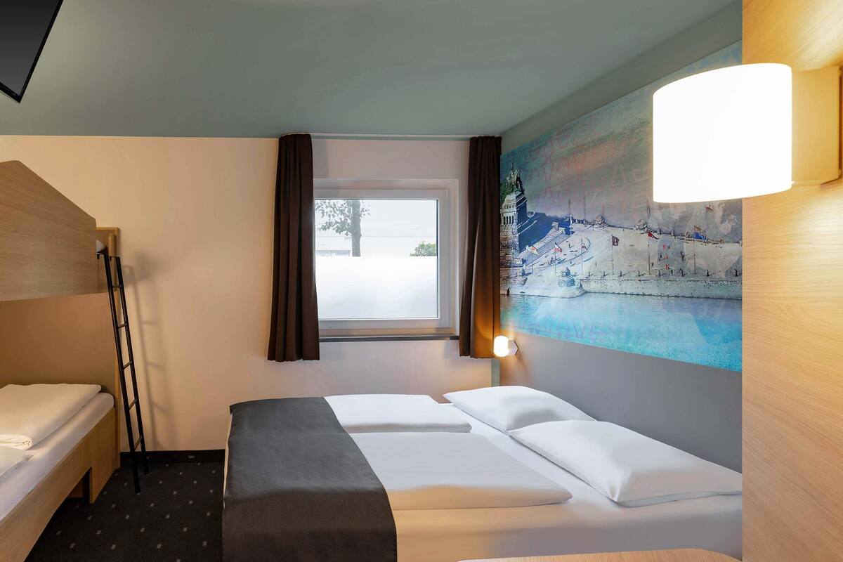 A hotel room with a bunk bed, a single bed, a bench, a window with curtains, and a wall-mounted lamp.