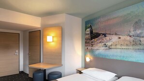 A hotel room with two beds, a small table, and a wall-mounted light. - B&B HOTEL Koblenz-City (Koblenz)