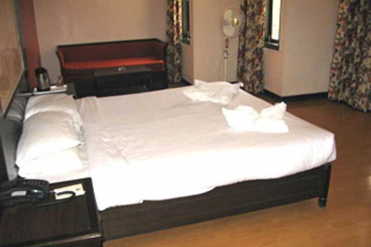 A neatly made bed with white linens and pillows, a wooden headboard, a bedside table with a lamp, and a floral curtain.