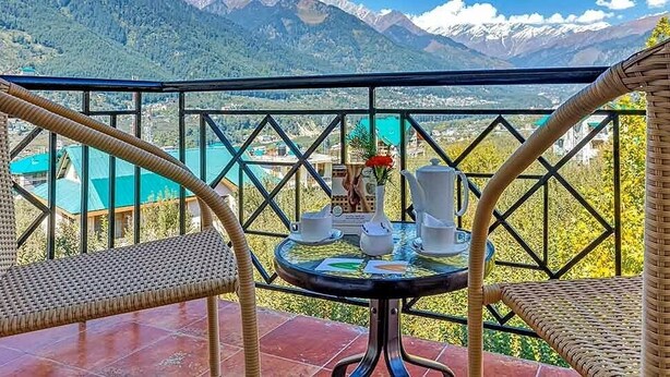 Un balcon avec des fauteuils en osier, une table ornée d’un vase et de tasses à thé, et une vue sur les montagnes.