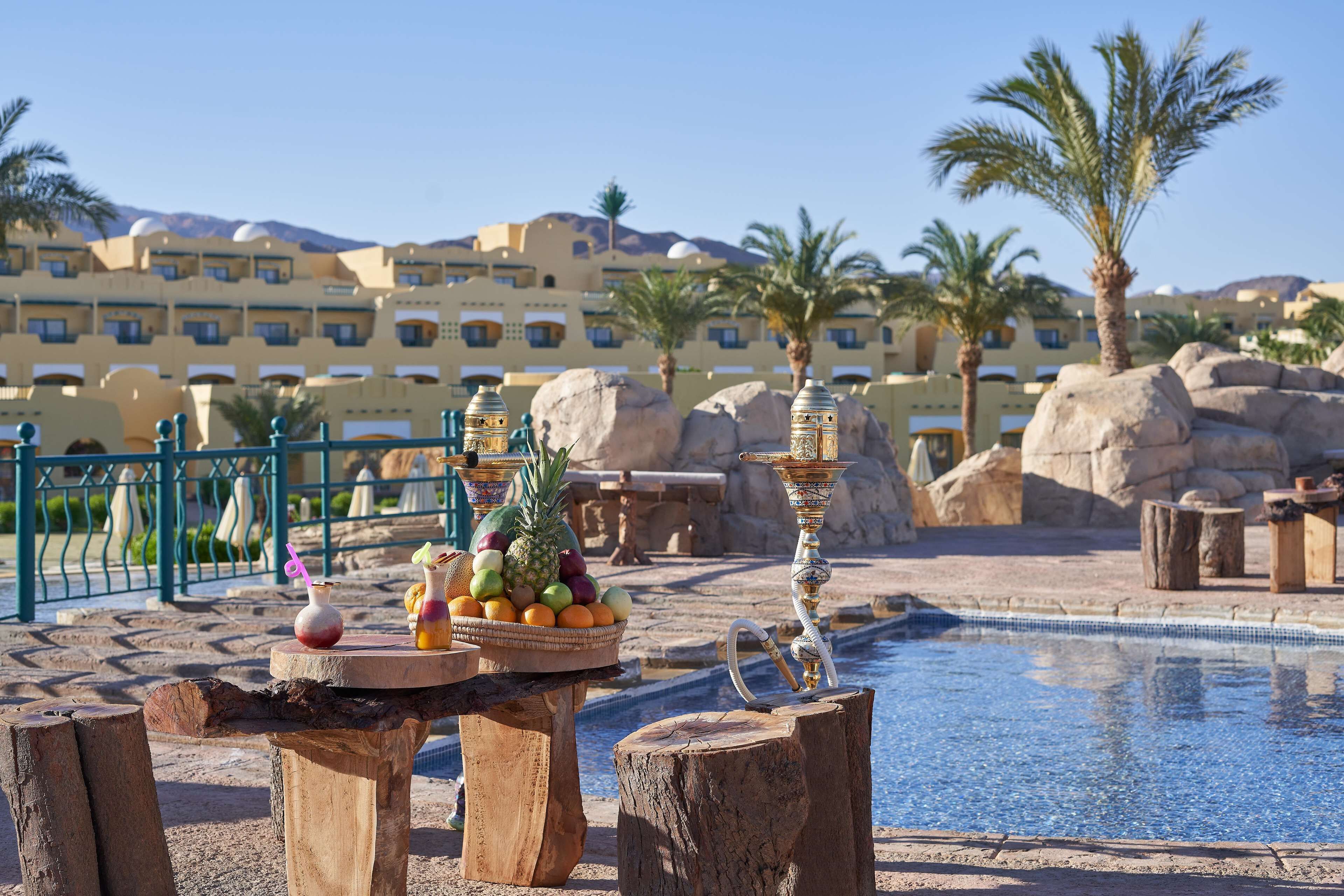 A pool area with a fruit bowl and decorative vases on wooden stools, surrounded by palm trees and large rocks.