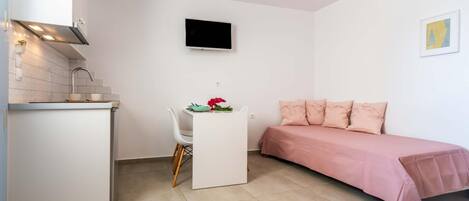Quarto compacto com cozinha compacta, pequena mesa de jantar, sofá e TV na parede.