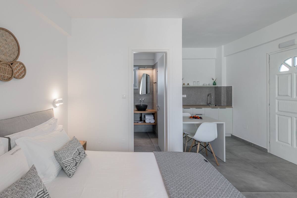 Quarto moderno com uma cama grande, luminárias de cabeceira e vista para uma cozinha e banheiro integrados.