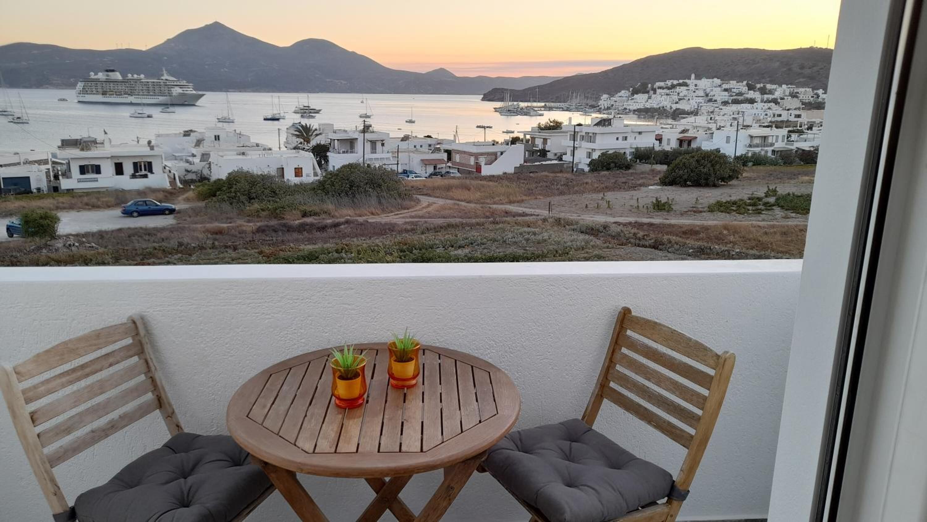 A balcony with a wooden table and two chairs, overlooking a coastal town with white buildings and boats in the water.